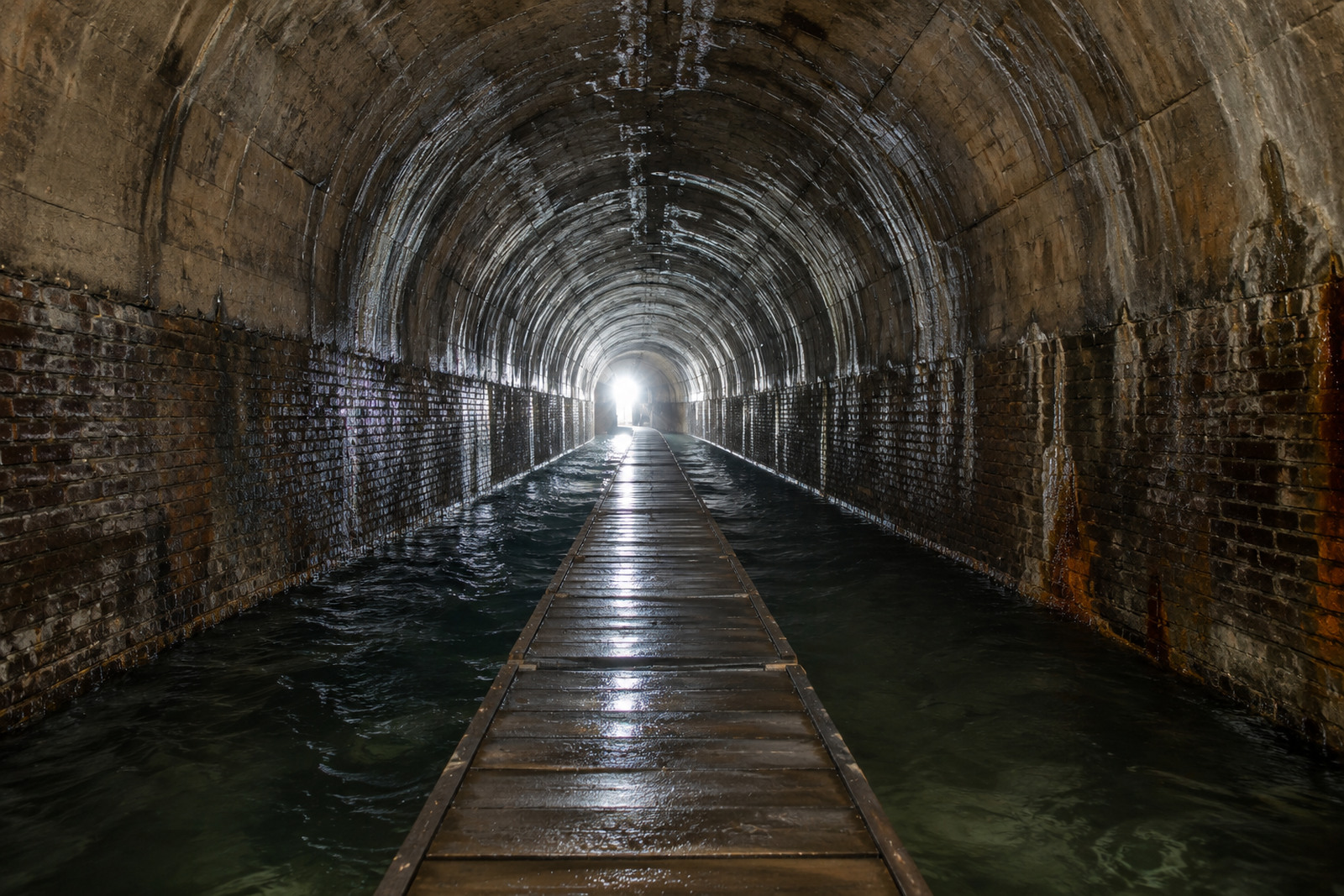 Long vaulted brick and concrete tunnel with a wooden walkway leading toward a bright opening at the far end